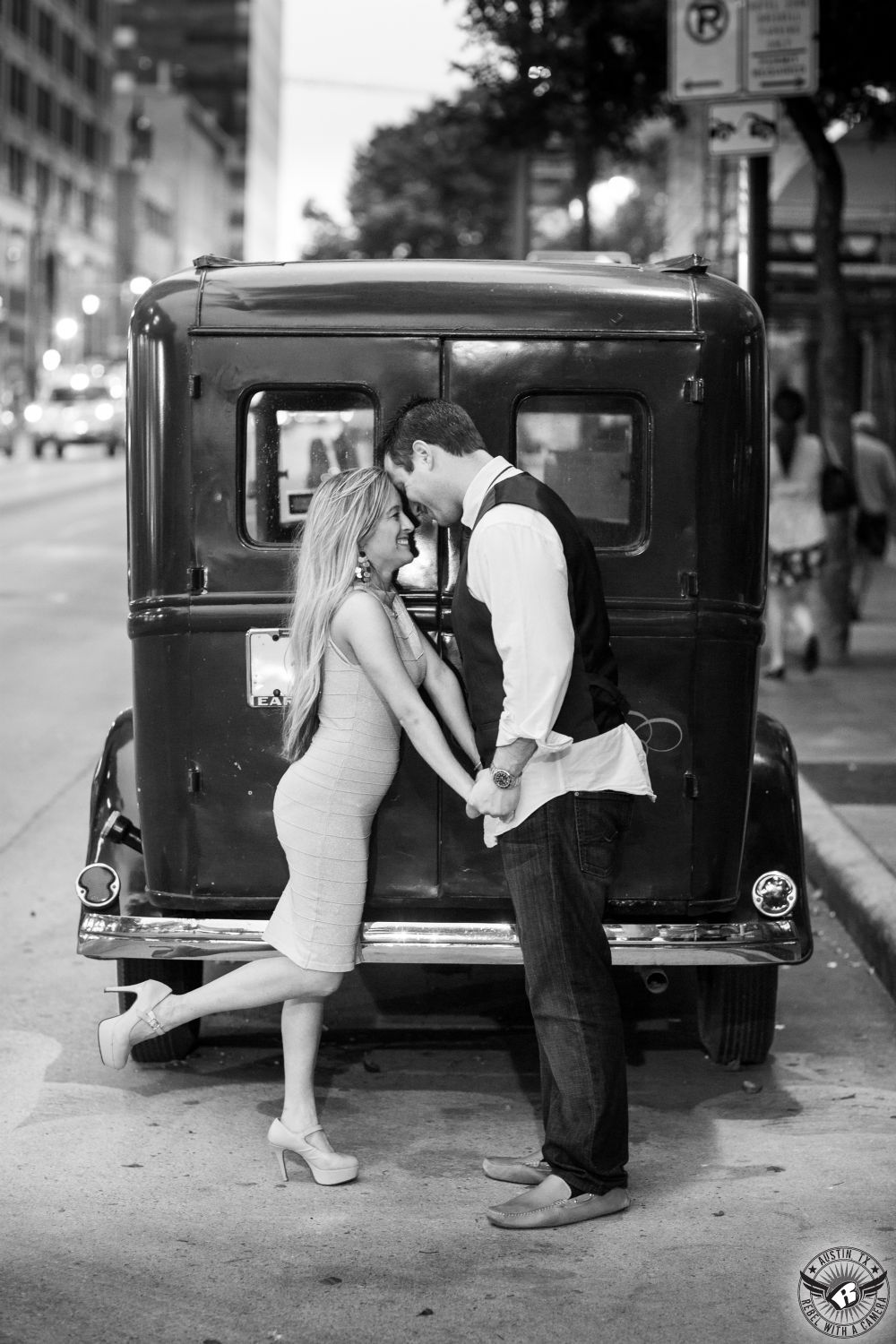 Blond, petite lady in a tight short light colored dress in flesh colored heels stands on one foot and holding hands with tall dark haired guy wearing untucked white dress shirt and dark blue jeans in front the rear doors of antique delivery truck on 6th street  near the Driskill Hotel in this adventurous engagement session in downtown Austin. 
